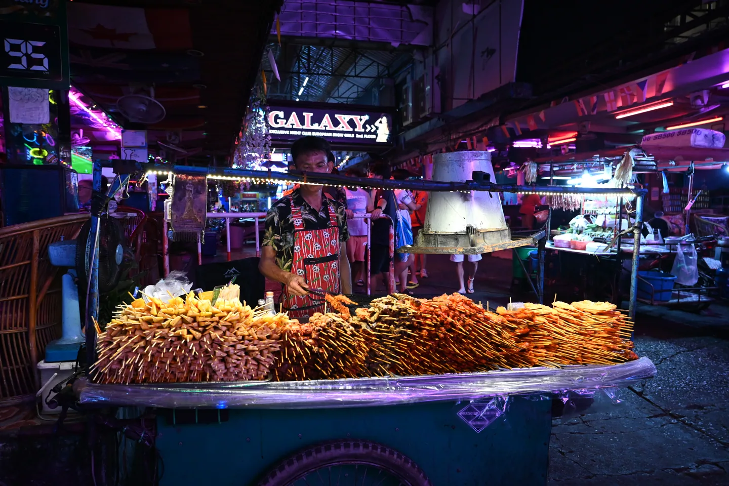 A vendor behind a cart of skewered food on a neon-lit Pattaya street.